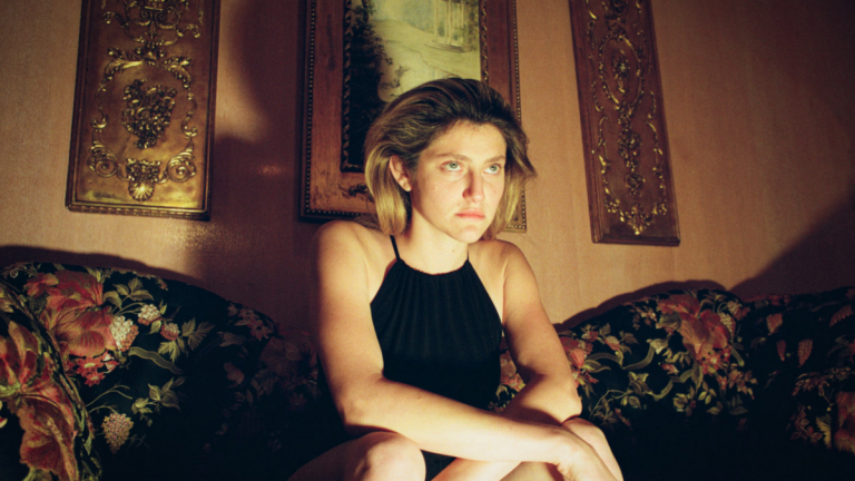 A woman wearing a black dress sits on a colorful couch in low lamp lighting with an ominous expression on her face.